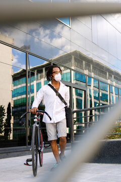 Man wearing face mask walking with bicycle against modern building during Covid-19 in city