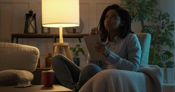 Thoughtful african american woman writing checklist in notepad late night at home