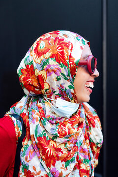 Cheerful woman in floral hijab against black wall during COVID-19