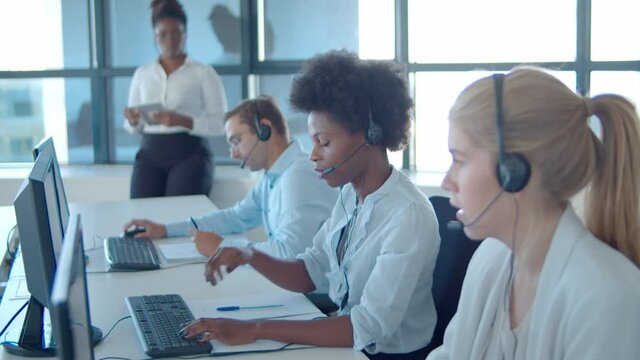 Female African American mentor watching new call center operators in headsets working at computers and consulting each others. Female business leader or telemarketing concept