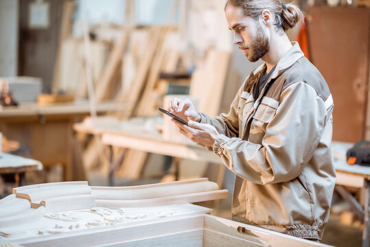 Handsome carpenter working on wooden carvings using a smart phone at the carpentry manufacturing. Concept of using mobile technologies in carpentry - Powered by Adobe