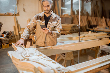 Handsome carpenter working on wooden carvings using a smart phone at the carpentry manufacturing....