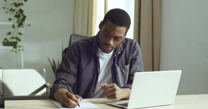 Portrait smart focused student black business man afro american guy sitting at table in home office looking at laptop screen making notes writes on paper doing test learning remotely studying language