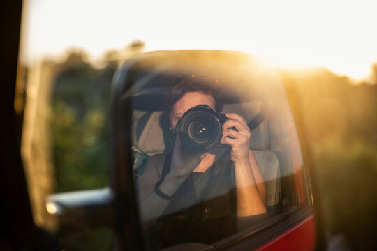Side-view mirror reflection of woman photographing setting sun