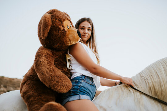 Portrait of beautiful young woman riding horse with large teddy bear behind