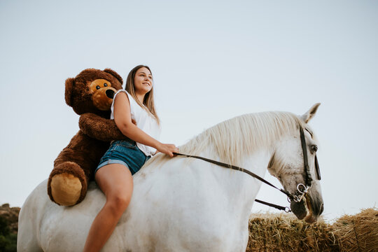 Beautiful young woman riding horse with large teddy bear behind