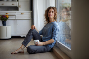 Mid adult woman with coffee cup sitting while leaning on glass door at home