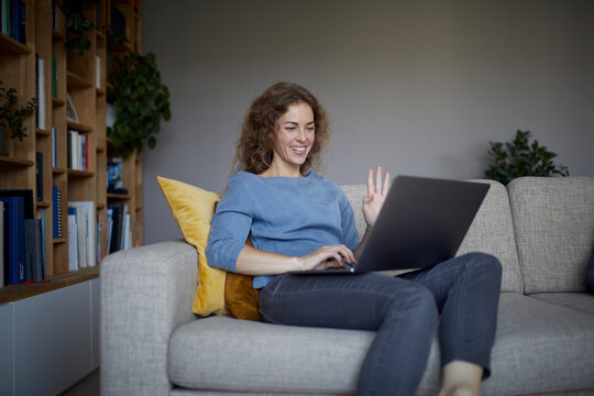 Mid Adult Woman Waving Hand To Video Call On Laptop While Sitting At Home