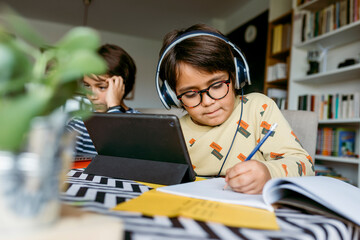 Boy writing notes through digital tablet sitting with male friend during e-Learning at home