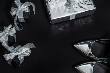Silver women's pumps, black gifts with silver ribbons with bows lie next to the gift wrapped in foil on black. View from above. Close up. Copy space