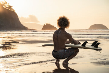 Shirtless young man with surfboard crouching at seashore during sunset