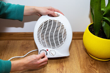 A girl holds her hands near a plastic fan heater and warms her hands, adjusting the temperature at...