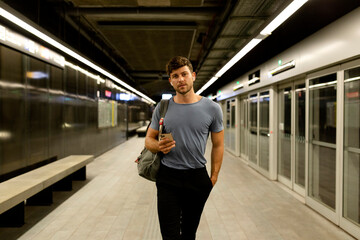 Handsome young man holding smart phone while walking with hand in pocket at illuminated subway platform