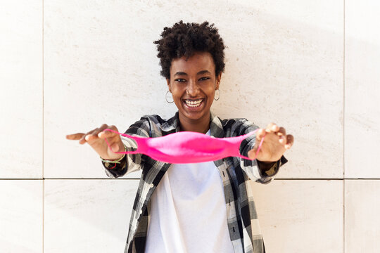 Cheerful young woman pulling pink mask against wall