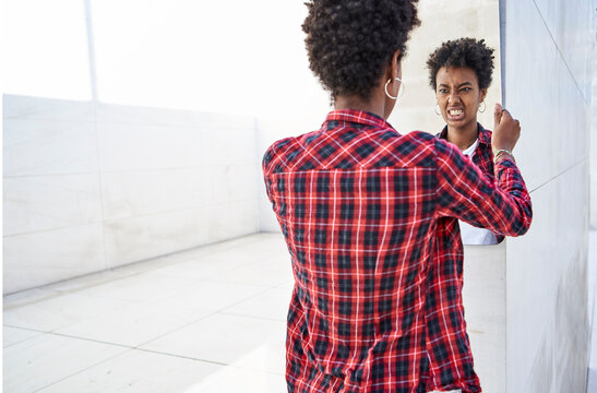 Young female hipster making a face while looking at mirror reflection against wall