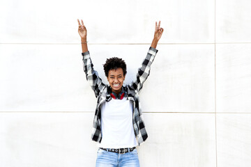 Cheerful female Afro hipster showing peace sign with arms raised against white wall