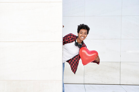 Happy young woman holding red heart shape balloon while standing behind white wall - Powered by Adobe