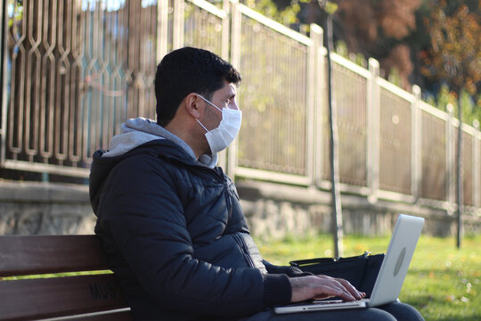 A young businessman with a protective face mask working on a laptop outdoors in the street. Social distancing during the coronavirus pandemic. Covid-19 epidemic in a city.