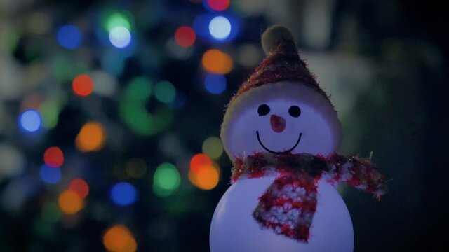 Snowman decoration with hat and scarf. Lights blink on Christmas tree in background with bokeh. Fun yard ornaments celebrate holiday in winter.