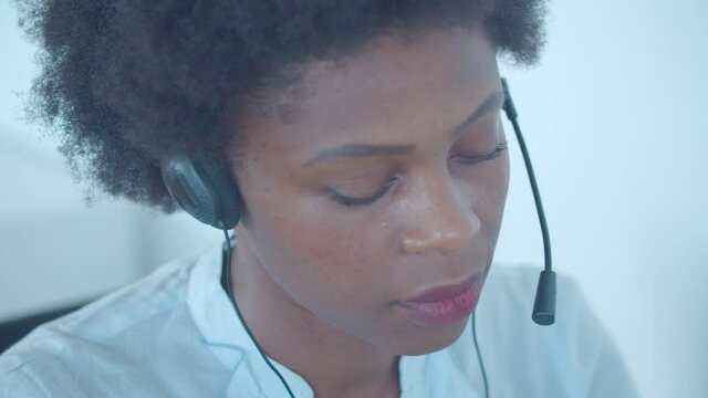 Focused African American employee in headset taking call, nodding and speaking at microphone at her workplace in call center. Closeup shot. Customers assistance or telemarketing concept