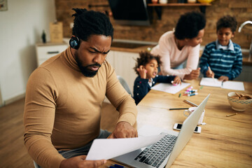 Black father using laptop and analyzing paperwork while working at home.