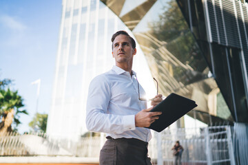 Pondering proud CEO in trendy outfit using touch pad application for creating web project during work break in financial district, Caucasian businessman thoughtful looking away sitting at urban street