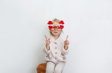 a little girl in funny Christmas glasses is sitting against a white wall.