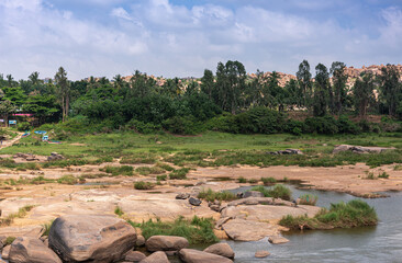 Hampi, Karnataka, India - November 4, 2013: Ferry business on opposite shore of Tungabhadra River just north of Viriupaksha Temple complex. Brown boulders, green forest, blue cloudscape, and yellow bo