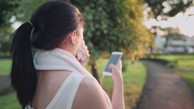 Over Shoulder Shot Asian Woman Using Smartphone While Walking Down On The Walking Path At Outdoor Garden During Warm Sunset Hour, Relaxing After Workout, Wiping Sweat From Face, Technology Lifestyle