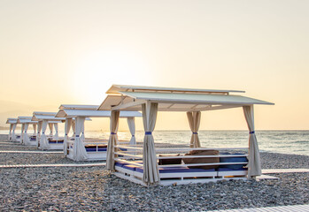Beach canopies with white curtains on a pebble beach by the sea at sunrise. Summer beach vacation and travel concept.