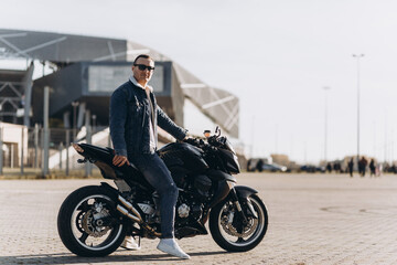 Fototapeta na wymiar A man in denim clothes sitting on a black sports motorcycle against the backdrop of a large modern building