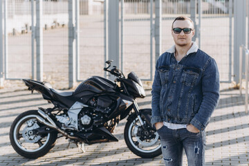 Naklejka na ściany i meble A man in denim clothes sitting on a black sports motorcycle against the backdrop of a large modern building
