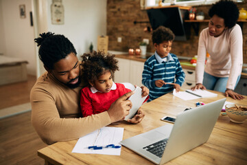 Small black girl sitting on father's lap while he is working at home.