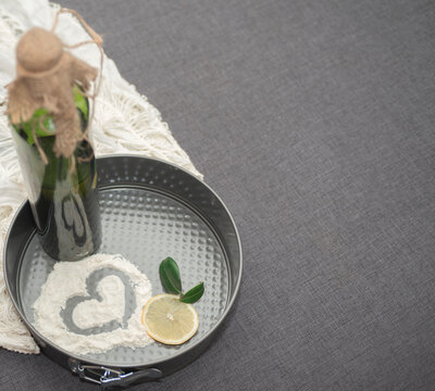 Composition of kitchen items on a gray background. Drawing on flour in the form of a heart on a metal cake mold. Nearby is a green bottle with olive oil and a slice of lemon with leaves