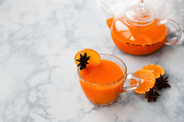 Sea buckthorn tea in a glass cup with orange and spices and a glass teapot on a light blue marble background.