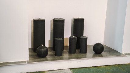 Black candles of various different shapes stand on the floor against the background of a white wall.
