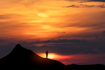 Beautiful dark golden sunset. Silhouette of a tourist on a hill stands and looks at the huge sun.