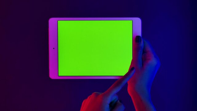 African American woman holds a white tablet in her hand and touches green screen chroma key. Close up gadget in horizontal position against a background of bright multicolored neon lights. Slow motion