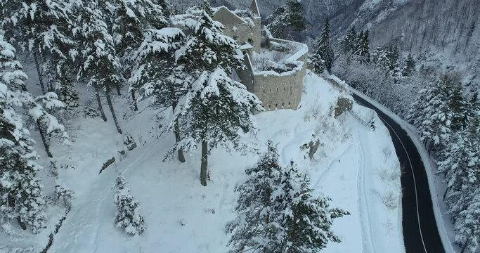 Aerial drone overhead view of medieval Predil fortress in Slovenia. Paved countryside road between forest. Cold winter in Alpine valley. Beautiful landscape covered with snow. Descending