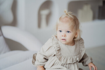 Fototapeta premium beautiful little girl of European appearance blonde with a ponytail. In a beige dress posing stands playing near the sofa and looks away. Bright and cozy apartment in the city center.