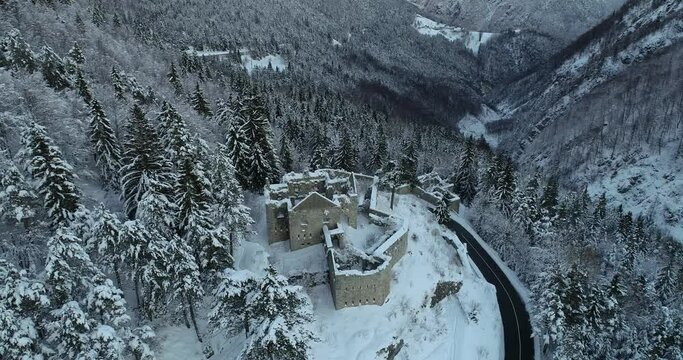 Aerial drone overhead view of medieval Predil fortress in Slovenia. Paved countryside road between forest. Cold winter in Alpine valley. Beautiful landscape covered with snow. Parallax moving