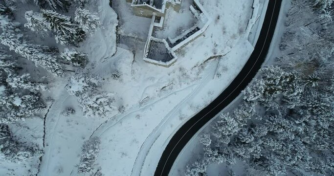 Aerial drone overhead view of medieval Predil fortress in Slovenia. Paved countryside road between forest. Cold winter season in Alpine valley. Beautiful landscape covered with snow. Forward moving