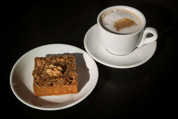 walnut cake on a white plate next to a Cup of cappuccino, Sunny photo. 