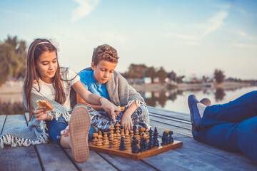 Two young cute little friends, boy and girl having fun while playing chess sitting by the lake in...