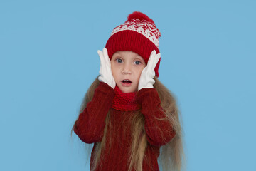 Winter portrait of happy child girl wearing knitted red hat, snood and sweater. Wow face. Blue background. childrens winter entertainment