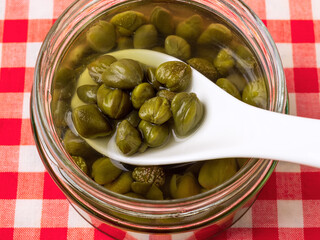 Taking pickled capers from a glass jar with white porcelain spoon close-up. Marinated buds of caper bush. Mediterranean cuisine ingredient. Organic spices and seasonings.