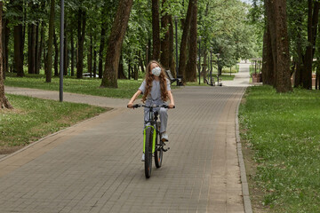 Obraz premium A girl in a medical mask rides a Bicycle in the Park in the summer along the path. Outdoor activity.