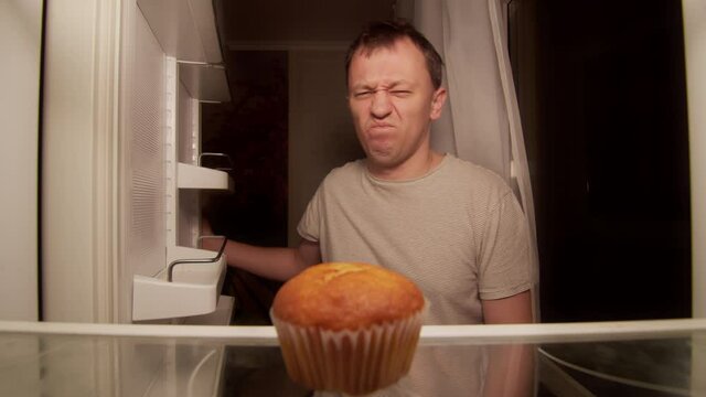 A man opens the refrigerator at night on the shelf is one cupcake, a disgruntled man