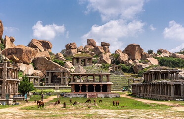 Hampi, Karnataka, India - November 4, 2013: Nandi Monolith Statue temple and nearby area. Cattle grazing in front. Ruinous buildings and big boulders on hill under blue cloudscape.