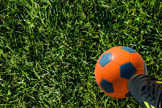 Futebol - relvado com uma bola cor de laranja e um pé de um jogador com uma bota pousado por cima da bola  - bola de cores laranja e azul - chuteira em verde fluorescente e preto